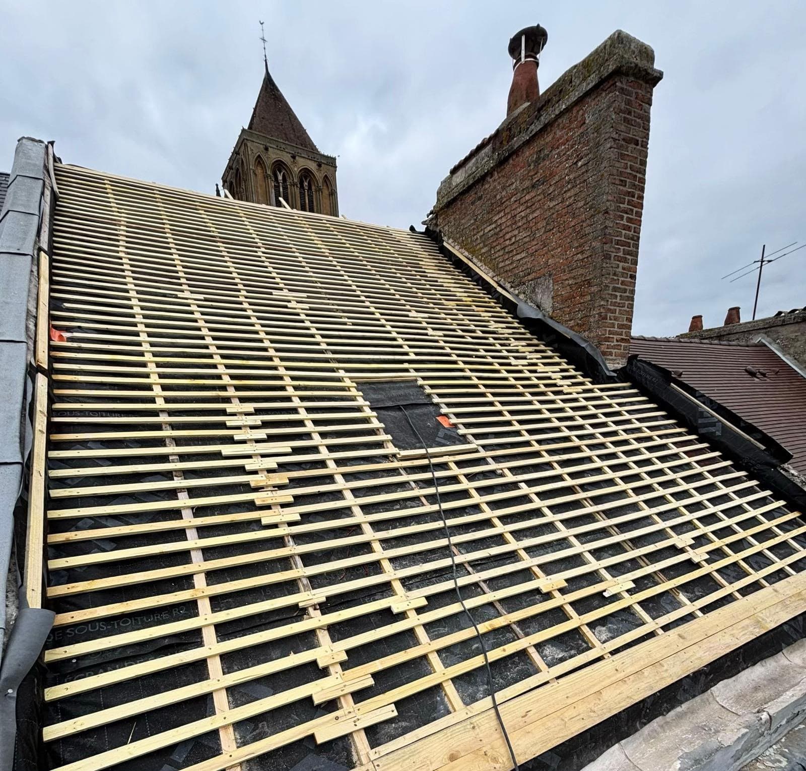 Lattes de bois sur une toiture en rénovation avec cheminée en briques et clocher d'église.